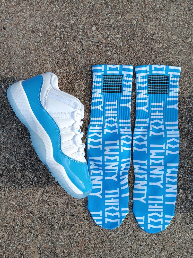 May include: A white and light blue trainer with a clear sole is next to two pairs of blue socks. The socks have the words "TWENTY THREE" in white. The trainer has a shiny light blue toe.