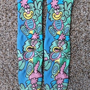 May include: A pair of blue, knee-high socks with a cartoon monster design. The monster is green, yellow, pink, and blue, with a peace sign. The socks have a blue background with green and pink accents.