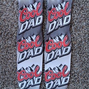 May include: A pair of grey and white socks with the words "Cool Dad" repeated on each sock. The socks feature a mountain graphic and a red and white colour scheme.