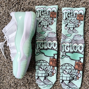 Puede incluir: Una zapatilla deportiva verde menta y blanca junto a dos pares de calcetines verde menta. Los calcetines presentan un personaje de dibujos animados de un iglú que sostiene un cartel que dice "IGLOO 11S SOLD HERE".
