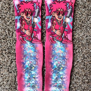 以下が含まれることがあります： ピンク色の髪、サングラス、ギターを持った漫画のキャラクターが描かれたピンク色の靴下のペア。デザインにはキラキラした紙吹雪が含まれています。靴下は明るく印象的な外観で、どんな服装にも彩りを添えるのに最適です。