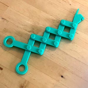 Könnte beinhalten: Ein türkisfarbenes, bewegliches Spielzeug in Form einer Hand mit einem Zeigefinger. Das Spielzeug besteht aus verbundenen rechteckigen Teilen und kreisförmigen Gelenken. Das Design umfasst zwei kreisförmige Öffnungen und eine Hand mit einem Zeigefinger.