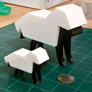 Peut inclure: Deux figurines de moutons imprimées en 3D, l'une plus grande que l'autre. Les moutons sont blancs avec des pattes et des visages noirs. Les moutons sont sur un tapis de coupe vert.