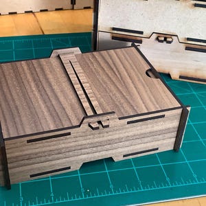 Puede incluir: Una caja de madera con tapa abatible y cierre. La caja está hecha de madera oscura con un patrón de veta natural. La tapa está cerrada y el cierre está en posición cerrada.