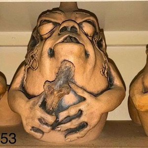 May include: Three ceramic jugs with human-like features. The jugs are in shades of brown and tan, with detailed facial expressions and hand-like appendages. The central jug holds an object. The number "53" is visible on the shelf below the jugs.