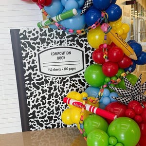 May include: A large composition book cutout with black and white speckled cover and lined paper design, accompanied by a vibrant balloon arch featuring red, blue, green, and yellow balloons, a yellow pencil balloon, and red and green marker balloons.