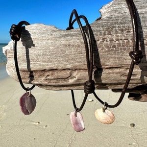 May include: Three leather necklaces with shell pendants. The necklaces are brown leather and the pendants are a pink and white shell, a purple shell, and a white shell.