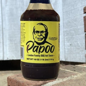 May include: A yellow and black bottle of Papoo London Family BBQ hot sauce. The bottle has a picture of a man with glasses and the text "Homemade Papoo" on it. The bottle is 18 ounces (1 lb 2 oz) or 510 grams.