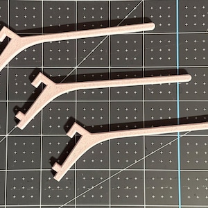 May include: Three light pink, curved plastic mask extenders are arranged on a dark gray cutting mat. Each extender has a hook on one end and a slot on the other. The extenders are designed to relieve ear strain from face masks.