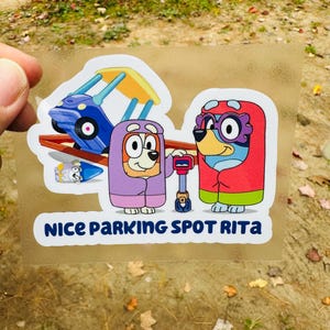 May include: A colorful sticker featuring cartoon characters in hooded outfits. The sticker includes the text "NICE PARKING SPOT RITA". The characters are in front of a blue and yellow playground structure.