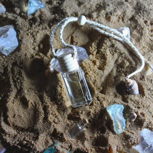May include: A clear glass bottle with a white cap and a string hanging from it. The bottle is filled with a clear liquid and is sitting on a bed of sand. There are some pieces of broken glass and a seashell in the sand.