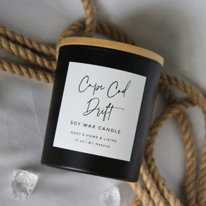 May include: Black soy wax candle with a wooden lid and a white label that reads "Cape Cod Drift" Soy Wax Candle Root's Home & Living 10 oz | All Natural