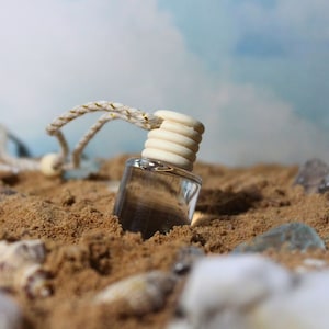 Pode incluir: Uma garrafa de vidro transparente com uma tampa de madeira e um cordão branco pendurado nela. A garrafa está parcialmente submersa na areia.