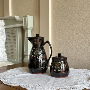 May include: A set of two brown ceramic containers with floral designs. The larger container is a jug with a spout and handle. The smaller container is a jar with a lid. Both containers have a dark brown glaze with gold accents and colourful floral designs.