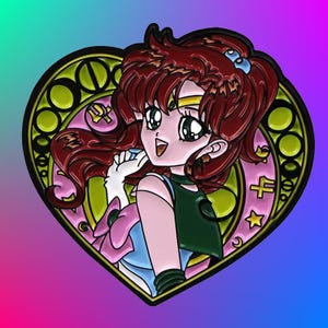 May include: A heart-shaped enamel pin featuring a cartoon character with long red hair, a green and blue outfit, and a pink and blue bow. The character is surrounded by a gold and green decorative frame.