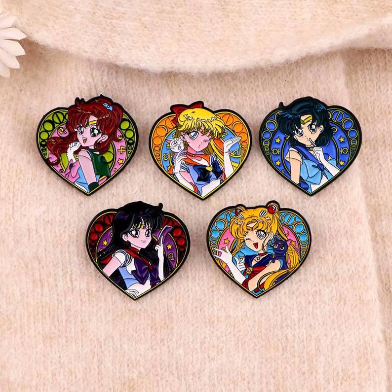 May include: Five enamel pins featuring characters from the anime Sailor Moon. Each pin is shaped like a heart and features a different Sailor Scout in a colorful, detailed design.