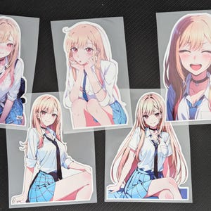 Puede incluir: Cinco pegatinas de estilo anime con un personaje rubio con uniforme escolar. El personaje se representa en varias poses, con una camisa blanca, una falda azul y una corbata negra. Cada pegatina está sobre un respaldo transparente.
