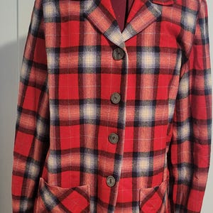 May include: A long-sleeve blazer featuring a red, black, and cream plaid pattern. The garment has a notched lapel, four dark buttons down the center front, and two square patch pockets at the lower front. The fabric has a soft, brushed texture.