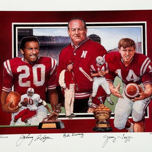 May include: A framed print featuring three football players in red and white uniforms, with a coach in a red jacket. The print includes signatures and a trophy. The background shows a stadium.