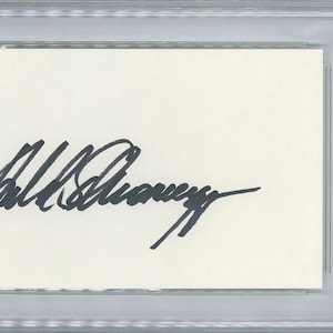 May include: A signed autograph on a cream-colored card, encased in a protective holder. The signature, in black ink, is of A. Schwarzenegger. A red and white label on the right side indicates authentication and certification.