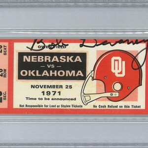 May include: A vintage ticket from the Nebraska vs. Oklahoma football game on November 25, 1971, featuring a red and white helmet graphic. The ticket is encased in a protective holder and includes the signature of Bob Devaney.