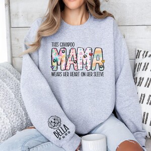 May include: A light gray sweatshirt with the text "THIS CAVAPOO MAMA WEARS HER HEART ON HER SLEEVE" in colorful letters. The left sleeve features a dog illustration and the name "BELLA". The design includes paw prints and hearts.
