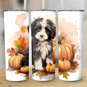 Puede incluir: Un vaso blanco con un diseño de acuarela de un perro blanco y negro, calabazas y hojas de otoño. El diseño envuelve el vaso, creando un tema de temporada. El vaso tiene una tapa plateada y una pajita.
