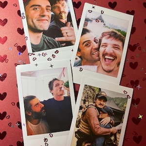 May include: Four instant film photographs featuring two men in various candid poses, including smiling, laughing, and embracing. The photos are arranged on a red surface decorated with scattered red heart-shaped confetti and small star-shaped accents.