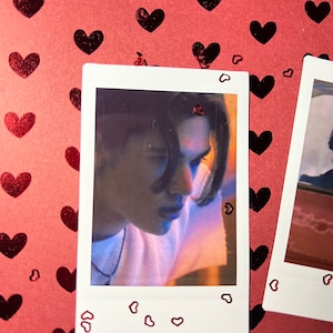 May include: A Polaroid photo of a person with long dark hair looking down. The photo is surrounded by a white border with small red hearts.