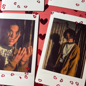 May include: Two Polaroid photos of a person with dark hair. The photos are on a red background with red glitter hearts. The person in the photos is wearing a striped shirt in one photo and a brown jacket in the other.