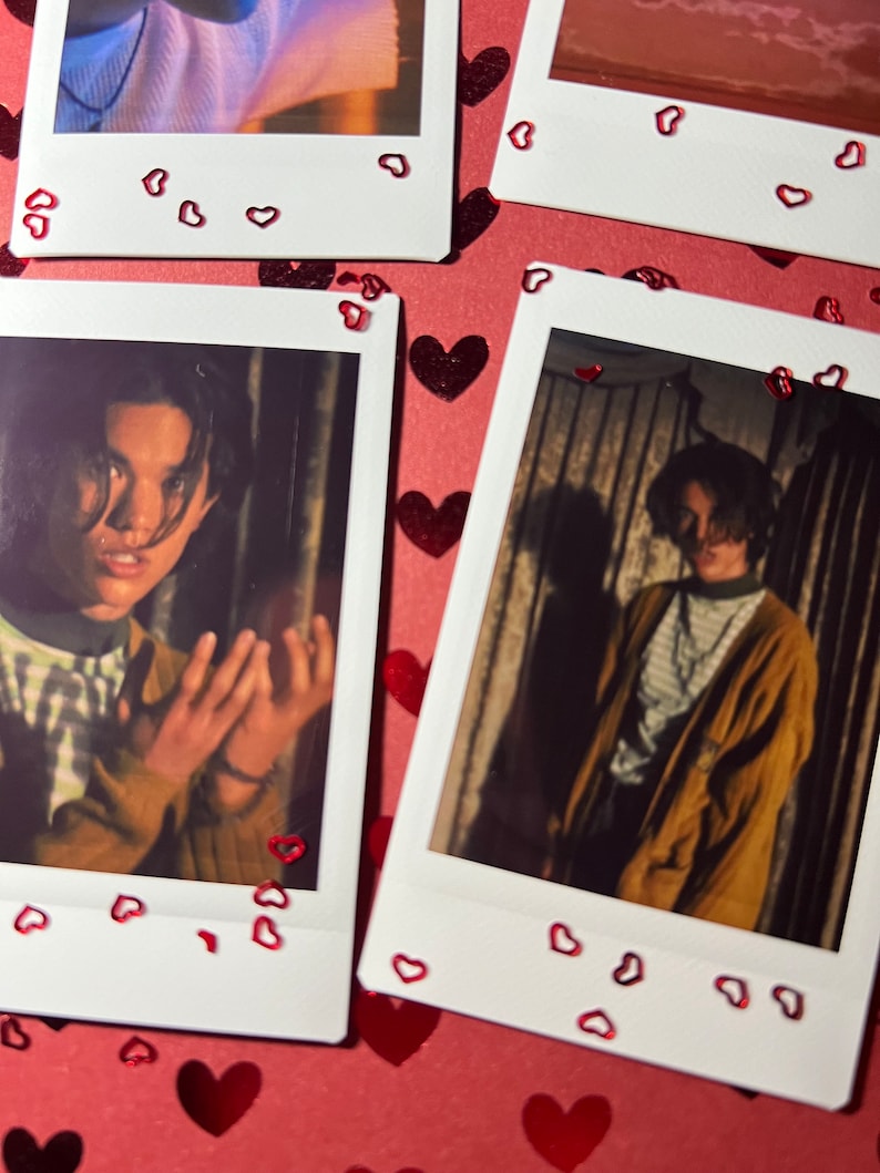 May include: Two Polaroid photos of a person with dark hair. The photos are on a red background with red glitter hearts. The person in the photos is wearing a striped shirt in one photo and a brown jacket in the other.