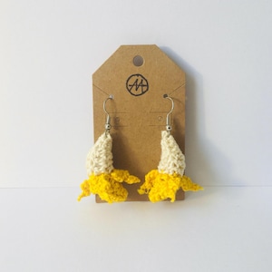 Puede incluir: Un par de pendientes de plátano hechos a mano con ganchillo. Los pendientes presentan una cáscara de plátano amarilla y un plátano de color crema. Los pendientes están sujetos a ganchos plateados y cuelgan de una etiqueta marrón con un logotipo.