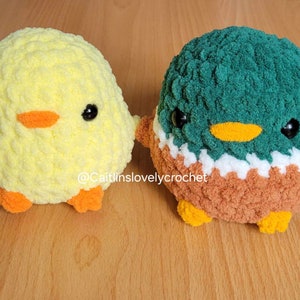 May include: Two plush, round, crocheted bird toys. One is yellow with an orange beak and feet, the other is green, white, brown, and orange with a yellow beak. Both have large black eyes. The text "@Caitlinslovelycrochet" is visible.