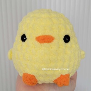 May include: A handmade, yellow crochet chick with black eyes and an orange beak and feet. The plush toy is round and fluffy, with a textured surface. The text "@Caitlinslovelycrochet" is visible on the bottom right.