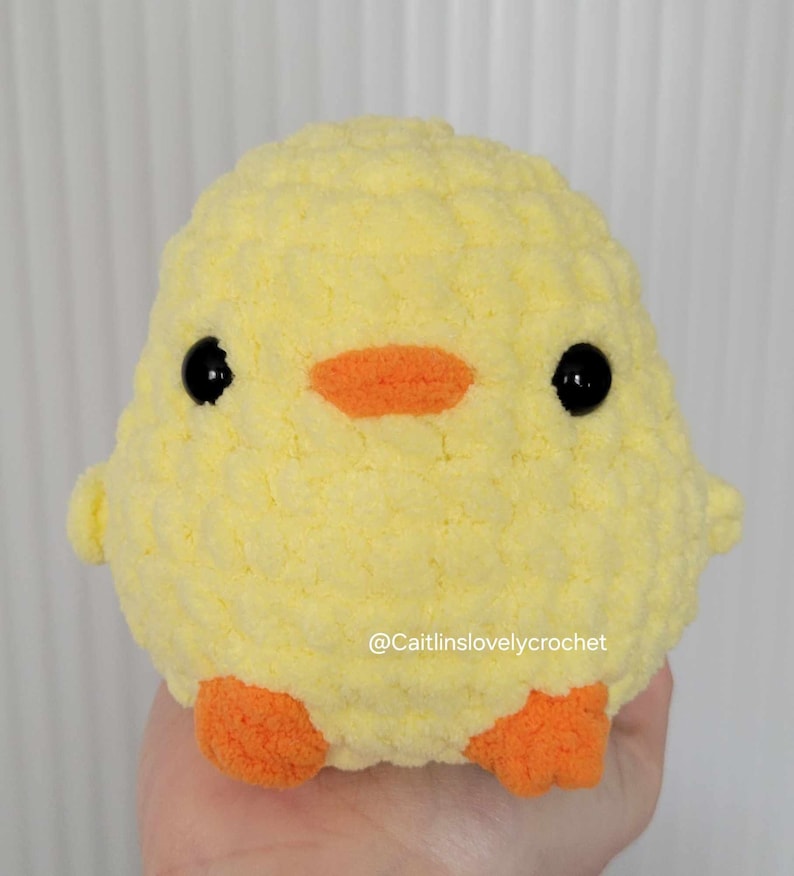 May include: A handmade, yellow crochet chick with black eyes and an orange beak and feet. The plush toy is round and fluffy, with a textured surface. The text "@Caitlinslovelycrochet" is visible on the bottom right.