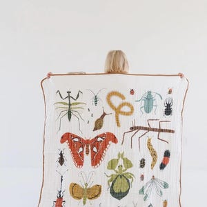 May include: White muslin blanket with a brown border, decorated with colourful illustrations of insects such as butterflies, beetles, and grasshoppers. The blanket is held up against a white background.