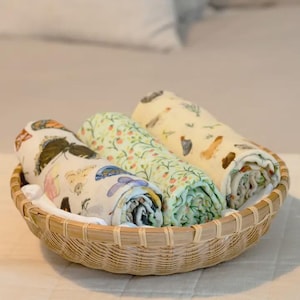 May include: A woven basket holds three rolled-up muslin blankets. The blankets feature colorful patterns of butterflies, flowers, and birds. The basket is made of light brown wicker and sits on a white surface.