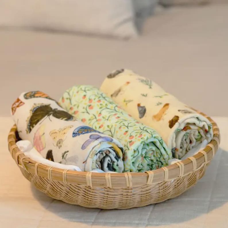 May include: A woven basket holds three rolled-up muslin blankets. The blankets feature colorful patterns of butterflies, flowers, and birds. The basket is made of light brown wicker and sits on a white surface.