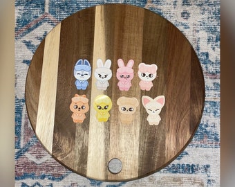 May include: A collection of nine cartoon character stickers arranged on a round wooden cutting board. The stickers feature various animal designs, including bunnies, bears, and foxes, in pastel colors. The board has a natural wood grain pattern.
