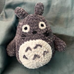 Puede incluir: Un juguete de peluche gris y blanco tejido a crochet que se asemeja a un personaje de dibujos animados con ojos grandes y un cuerpo redondeado. El juguete tiene una barriga blanca con un patrón de líneas curvas.