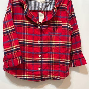 May include: Red plaid button-down shirt with long sleeves. The shirt features a classic plaid pattern in red, blue, yellow, and white. It has a chest pocket and a collar with a striped lining. The brand tag reads "Abercrombie & Fitch".