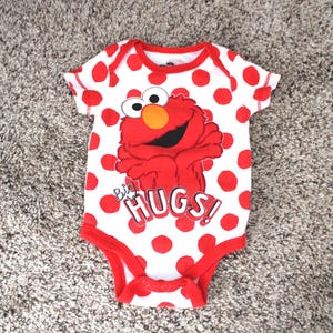 Könnte beinhalten: Ein weißer Baby-Body mit roten Punkten und roter Einfassung. Der Body zeigt eine Grafik von Elmo aus der Sesamstraße mit den Worten "BIG HUGS!" in einer verspielten Schriftart. Der Body ist für Säuglinge konzipiert.