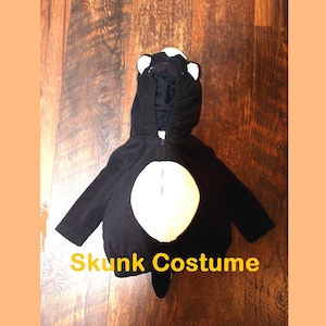 May include: A black skunk costume with a white belly and a hood featuring white ear accents. The costume has a zipper closure and a tail. The text "Skunk Costume" is displayed in yellow.
