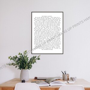 May include: A framed black and white print of "The Stranger" by Albert Camus, with text in a typewriter-style font. The print is on a wooden table, with a vase of greenery. The frame is black.