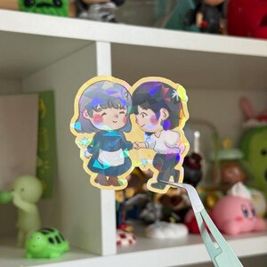 May include: A holographic sticker featuring two chibi-style characters holding hands. The character on the left has short gray hair and a blue dress with a white apron, while the character on the right has dark hair and a white shirt with dark pants.