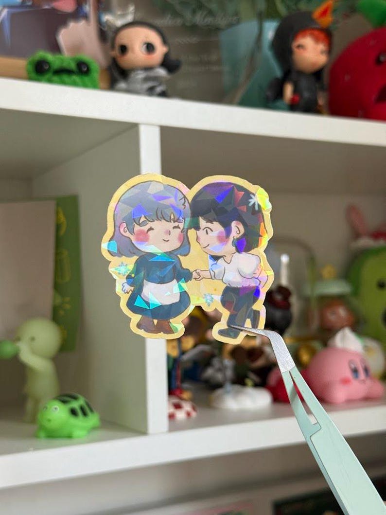 May include: A holographic sticker featuring two chibi-style characters holding hands. The character on the left has short gray hair and a blue dress with a white apron, while the character on the right has dark hair and a white shirt with dark pants.