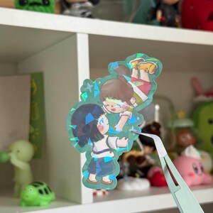 May include: A holographic die-cut sticker featuring an illustration of two anime-style characters reaching toward each other. The sticker has a shimmering, multi-faceted finish with blue and green light reflections across the surface of the artwork.