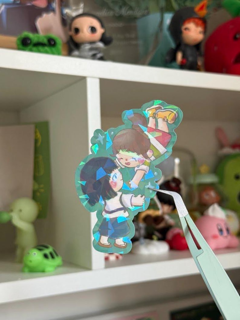 May include: A holographic die-cut sticker featuring an illustration of two anime-style characters reaching toward each other. The sticker has a shimmering, multi-faceted finish with blue and green light reflections across the surface of the artwork.