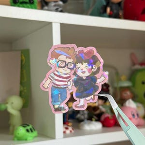 May include: A holographic sticker featuring a cartoon illustration of two characters holding hands. One character wears a red and white striped shirt with blue pants and glasses, while the other wears a dark dress with a red bow in their hair.
