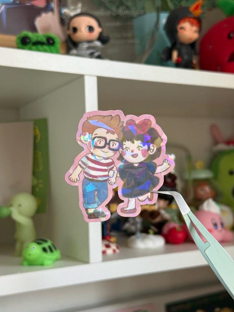 May include: A holographic sticker featuring a cartoon illustration of two characters holding hands. One character wears a red and white striped shirt with blue pants and glasses, while the other wears a dark dress with a red bow in their hair.